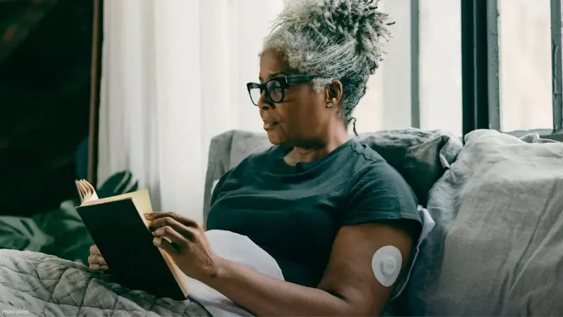 Senior woman with gray hair reading a book indoors wearing a glucose monitoring sensor., tags: diabetes - pexel