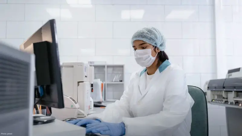 Scientist using a computer in a laboratory with protective gear including gloves, mask, and hair net., tags: healthcare - pexel